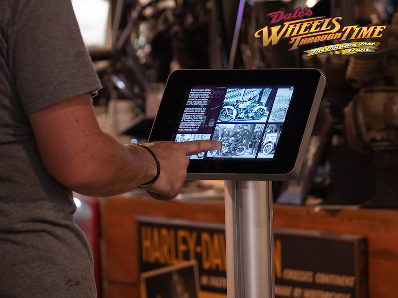 Visitor using an interactive iPad kiosk on a floor stand at the Wheels Through Time motorcycle museum
